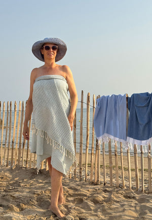 Frau steht am Strand mit dem Hamamtuch in Waffelpique Muster in Blau/Hellblau um den Körper gewickelt und schaut lächelnd in die Kamera